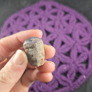 May include: A small, rough-textured, gray and brown stone with a smooth, dark blue top. The stone is held in a hand, with a purple crocheted background.