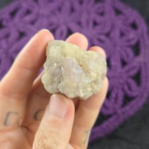 May include: A rough, light-colored stone with a mix of beige and pale purple hues. The stone is held in a hand against a blurred purple background, showcasing its natural texture and color variations. The stone appears to be a mineral specimen.