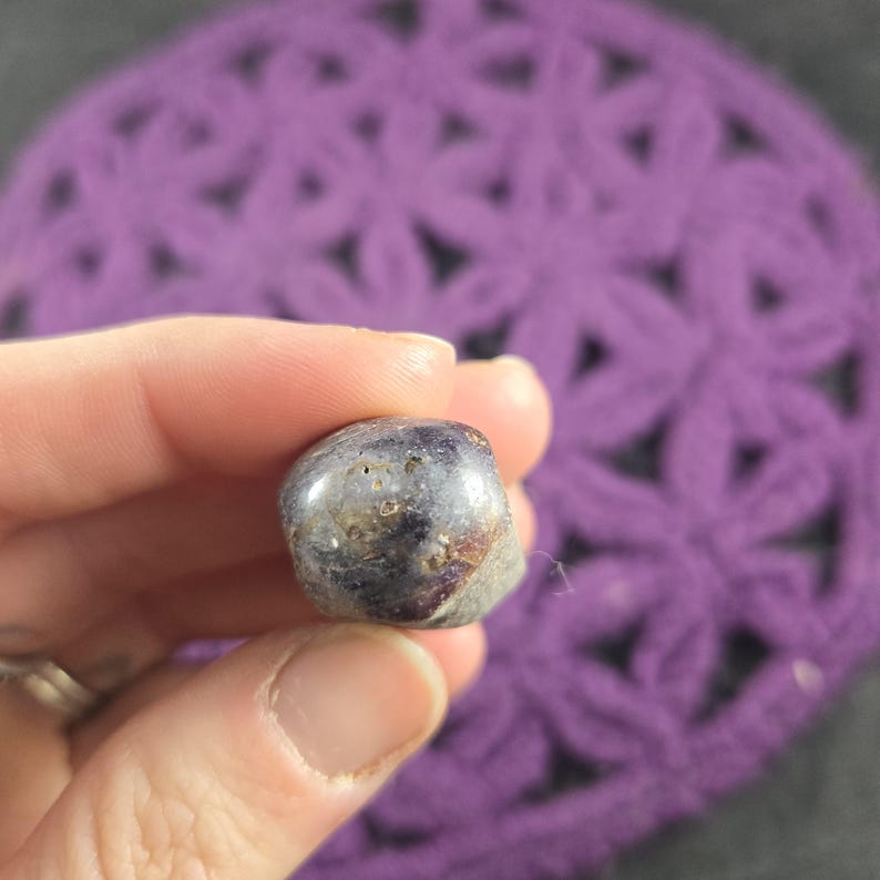 May include: A small, polished gemstone with a mix of dark purple, gray, and brown hues. The stone has a smooth, rounded shape and appears to be held in a hand. The background is a purple, crocheted surface.