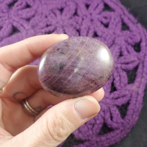 May include: A smooth, oval gemstone in shades of deep purple and burgundy, held in a hand. The stone has a polished surface with subtle veining and dark inclusions. The background is a textured, purple crocheted item.