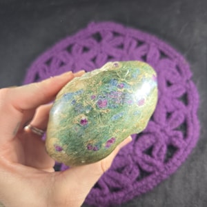 May include: A polished stone with a green, blue, and tan surface, speckled with small, vibrant red inclusions. The stone is held in a hand, with a purple crocheted background. The stone's natural colors and textures are highlighted.