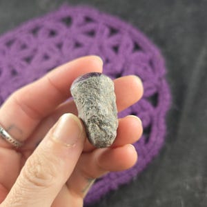 K&ouml;nnte beinhalten: Ein kleiner, rau strukturierter, grauer Stein mit einer polierten, dunkelvioletten Oberseite. Der Stein wird in einer Hand gehalten, mit einem lila geh&auml;kelten Hintergrund. Der Stein scheint ein nat&uuml;rlicher Edelstein oder ein Mineralienexemplar zu sein.