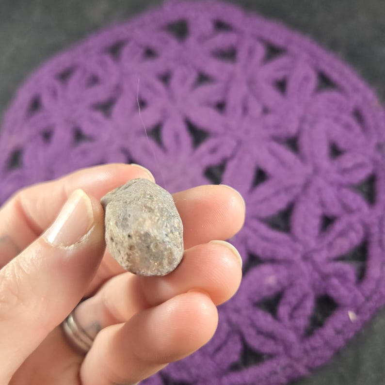 May include: A small, irregularly shaped, gray stone held in a hand. The stone has a rough, textured surface. A purple crocheted item with a floral pattern is in the background.