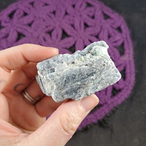 May include: A rough, gray and green crystal with a layered, striated texture. The crystal has a natural, raw appearance.