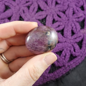 May include: A smooth, polished pink and purple gemstone with black and gold inclusions. The gemstone is round and has a slightly flattened shape.