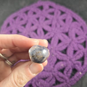 May include: A polished, rounded gemstone with a dark blue and brown color scheme. The stone is held in a hand, with a purple crocheted background. The gemstone has a smooth, reflective surface.