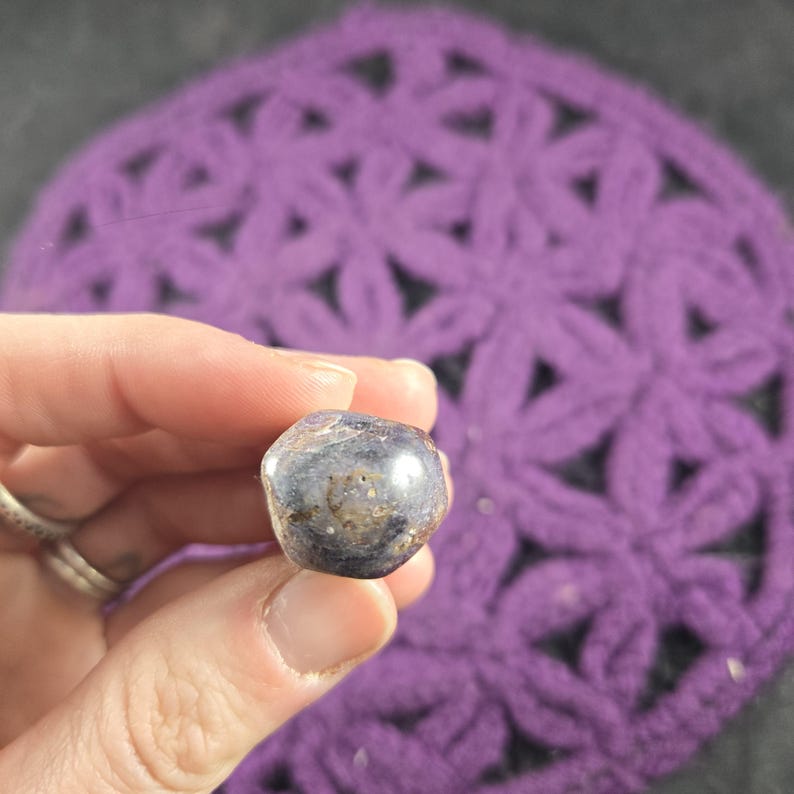 May include: A polished, rounded gemstone with a dark blue and brown color scheme. The stone is held in a hand, with a purple crocheted background. The gemstone has a smooth, reflective surface.