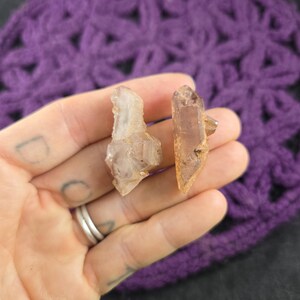 May include: Two raw, natural crystals are held in a hand. The crystals are light brown and beige, with a rough, textured surface. The background is a purple crocheted fabric.