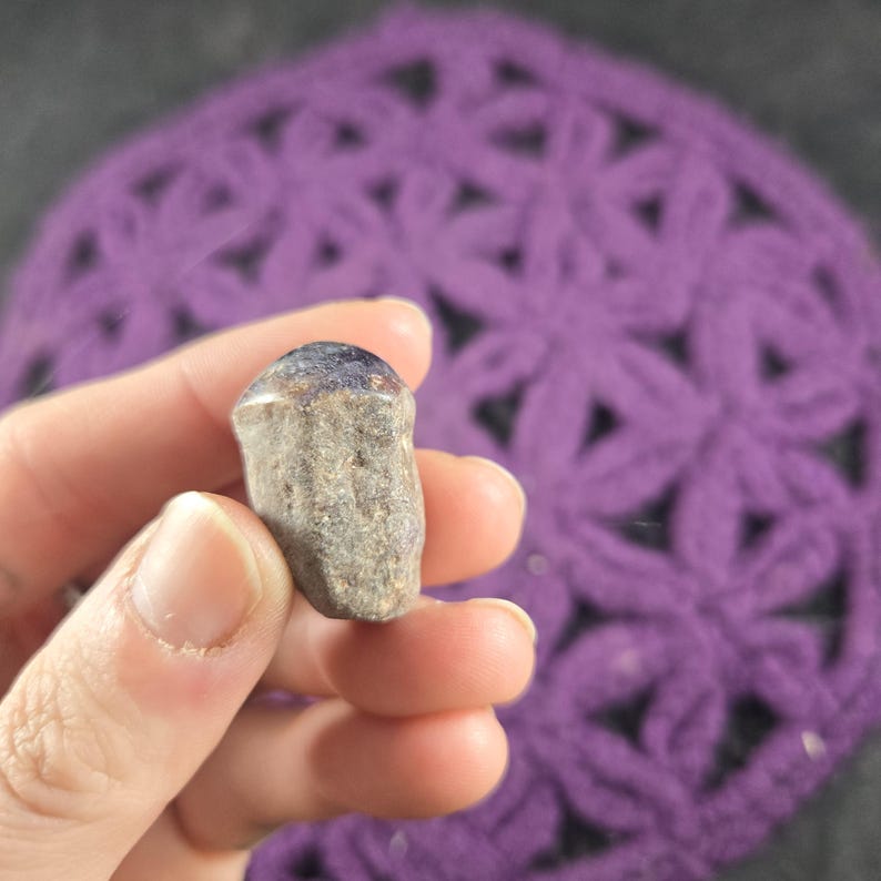 May include: A small, rough-textured, gray stone with a dark purple top is held in a hand. The stone is set against a blurred background of a purple crocheted doily.