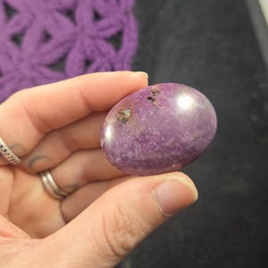 May include: A polished, oval-shaped purple gemstone with subtle marbling and small dark inclusions. The stone is held in a hand, highlighting its natural colour and texture. The background includes a purple crocheted item and a dark surface.