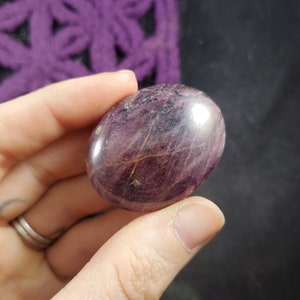 May include: A smooth, polished, oval-shaped purple gemstone with a dark red hue and white streaks. The stone is held in a hand.