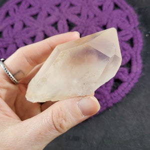 May include: A clear quartz crystal point with a double termination. The crystal is smooth and polished, with a slight cloudy appearance. The crystal is held in a hand, with a purple crocheted background.