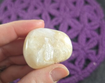 May include: A smooth, polished, cream-colored gemstone with subtle internal patterns. The stone is held in a hand, with a purple crocheted background. The gemstone has a rounded shape and a slightly translucent appearance.