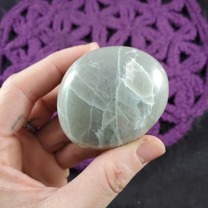 May include: A smooth, oval-shaped, pale green stone with white veining. The stone is held in a hand, and the background is a purple crocheted doily. The stone appears to be polished and has a glossy finish.