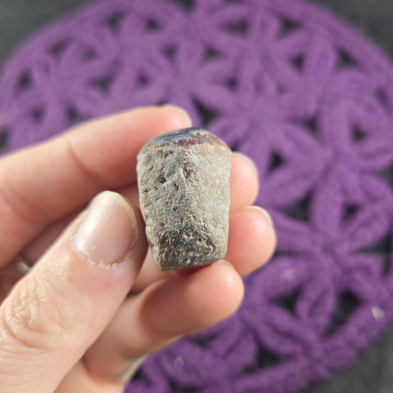 May include: A small, cylindrical bottle stopper with a textured, gray body and a dark blue and purple top. The stopper is held in a hand, with a purple crocheted background.