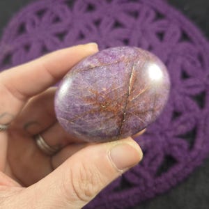 May include: A smooth, oval gemstone in shades of purple and brown, held in a hand. The stone has natural veining and a glossy finish. The background is a purple crocheted fabric.