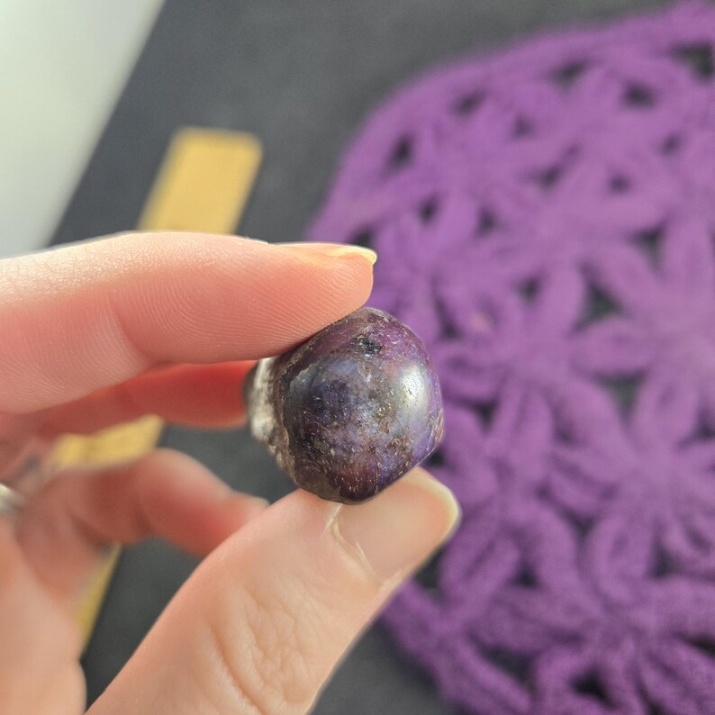 K&ouml;nnte beinhalten: Ein polierter, abgerundeter Edelstein mit tiefvioletter Farbe, gesprenkelt mit dunkleren Farbt&ouml;nen. Der Stein wird zwischen Fingern gehalten, was seine glatte, reflektierende Oberfl&auml;che zeigt. Der Hintergrund umfasst ein violettes H&auml;kelteil und eine dunkle Oberfl&auml;che.