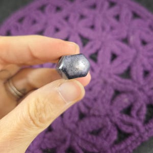 May include: A small, hexagonal, dark grey bead with a metallic sheen. The bead is held against a backdrop of a purple, crocheted fabric. The bead's surface reflects light, creating highlights.