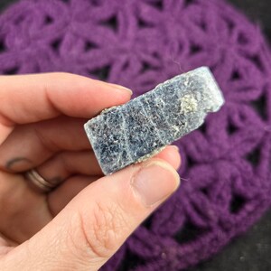 May include: A rough, gray-blue kyanite crystal with a rectangular shape. The crystal has a slightly iridescent sheen and is held in a hand.