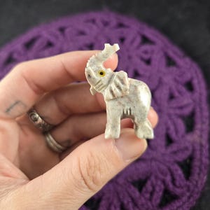 May include: A small, white stone elephant figurine with a yellow eye. The elephant is carved in a standing position with its trunk raised.