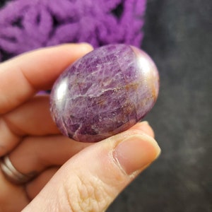 May include: A smooth, polished purple and brown gemstone with a rounded shape. The stone has a natural, striated texture.
