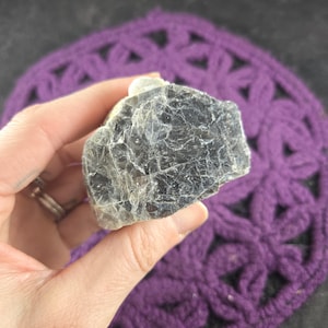 May include: A dark gray, rough-textured mineral specimen with light-colored veins. The stone is held against a purple, crocheted background. The mineral appears to be a natural, unpolished stone.
