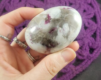 May include: A polished, oval-shaped gemstone with a mix of white, gray, and translucent areas, accented with deep pink and red inclusions. The stone is held in a hand, with a purple crocheted background.