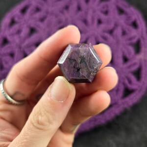 May include: A purple, faceted, star-shaped crystal with black inclusions. The crystal is being held in a hand.