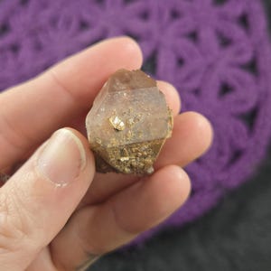 May include: A clear quartz crystal with a brown, rough exterior and small, gold-colored inclusions. The crystal is shaped like a pyramid and has a pointed tip.