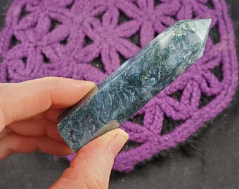 May include: A green moss agate crystal point with a pointed tip and a smooth, polished surface. The crystal is a dark green color with white and gray inclusions.