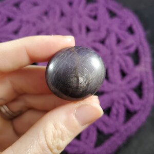 May include: A smooth, round, dark grey gemstone with a subtle shimmer. The gemstone is polished and has a reflective surface.