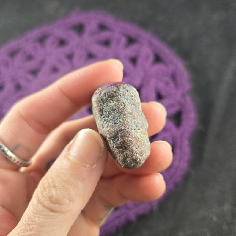 K&ouml;nnte beinhalten: Ein kleiner, l&auml;nglicher Stein mit rauer Textur, der eine Mischung aus Grau-, Braun- und Rott&ouml;nen aufweist. Der Stein wird in einer Hand gehalten, mit einem violetten H&auml;kelhintergrund. Die Oberfl&auml;che des Steins weist nat&uuml;rliche Unvollkommenheiten auf.