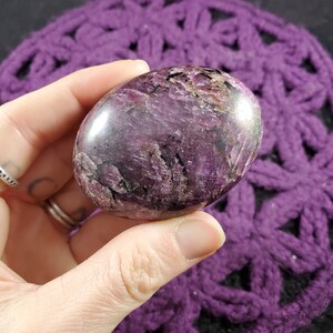 May include: A polished purple and black gemstone with a smooth, oval shape. The stone has a layered, striated pattern and is held in a hand.