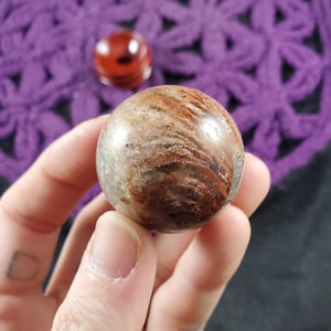 May include: A smooth, round, brown and white sphere with swirling patterns. The sphere is made of natural stone and is approximately 2 inches in diameter.