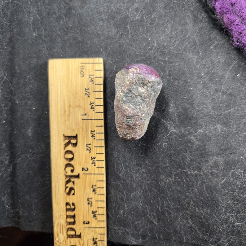 K&ouml;nnte beinhalten: Ein roher, nat&uuml;rlicher Edelstein mit einer tiefvioletten Oberseite und einem grauen und braunen K&ouml;rper. Der Stein liegt neben einem Holzlineal, das in Zentimetern markiert ist, mit dem Text "Rocks and" sichtbar. Der Hintergrund ist dunkel.