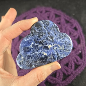 May include: A cloud-shaped sodalite stone with a striking blue and white marbled pattern. The polished gemstone is held in a hand, showcasing its natural beauty and unique veining. The stone is a decorative item.