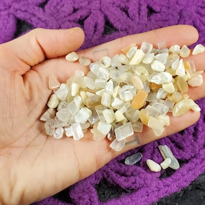 May include: A hand holding a pile of small, white, and peach-colored moonstone chips. The chips are irregular in shape and have a pearly sheen.