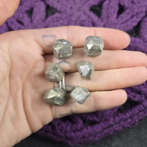 May include: Six pyrite stones, or fool's gold, are held in a hand. The stones have a metallic, silver-gold colour and a rough, faceted texture. The background is a purple, knitted fabric.