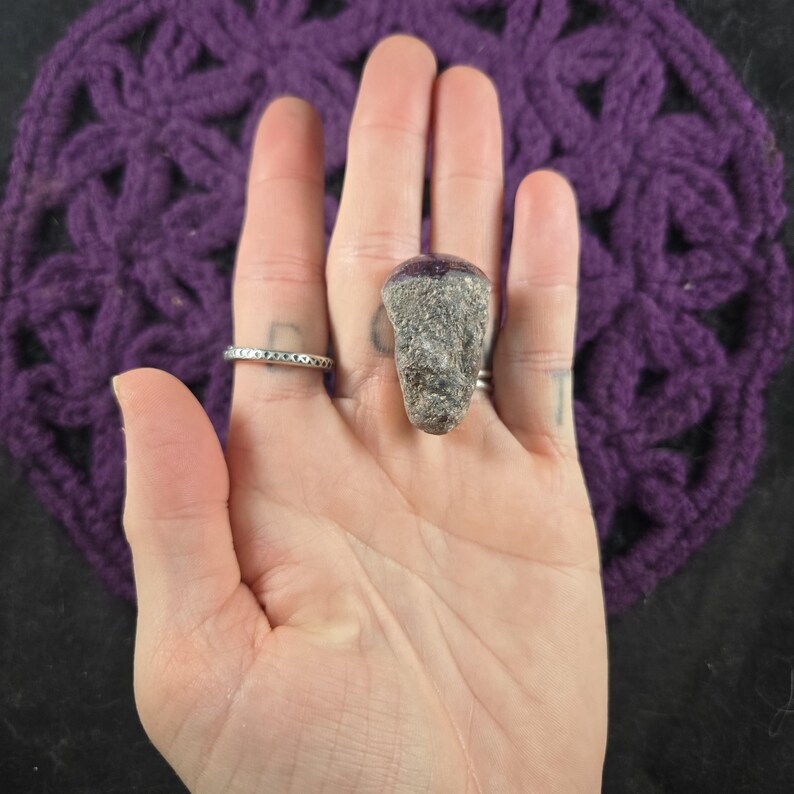 K&ouml;nnte beinhalten: Ein grauer Steinring mit strukturierter Oberfl&auml;che und dunkelvioletter Oberseite. Der Ring wird an einer Hand getragen, mit einem silbernen Bandring an einem angrenzenden Finger. Der Hintergrund zeigt ein lila H&auml;kelmuster.