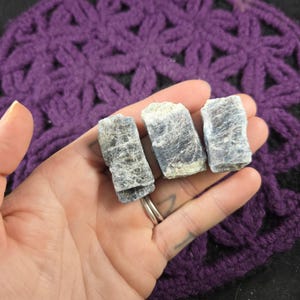 May include: Three rough, gray-blue kyanite crystals with a slightly iridescent sheen. The crystals are arranged in a row, with the largest crystal in the middle.