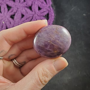 May include: A round, polished purple gemstone with a smooth, shiny surface. The stone has a light purple hue with darker purple streaks and a hint of gold.