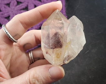 May include: A clear quartz crystal cluster with a double terminated point. The crystal has a natural, raw appearance with inclusions of brown and yellow minerals.