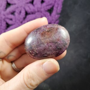 May include: A smooth, polished purple and red gemstone with a natural, irregular shape. The gemstone is held in a hand, showcasing its unique colour and texture.