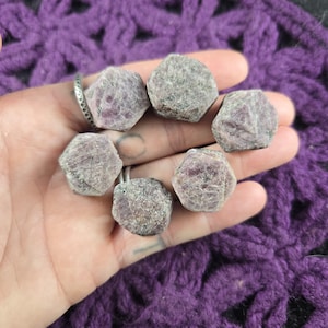 May include: Six rough-cut, hexagonal ruby gemstones in varying shades of pink and gray are held in a hand. The gemstones are arranged in a circular pattern. The background is a purple, crocheted fabric.
