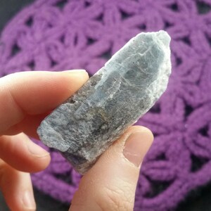 May include: A light blue and gray crystal with a rough, natural texture. The crystal is held in a hand against a purple background.