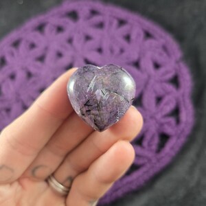 Peut inclure: Une pierre en forme de cœur, polie, présentant un mélange de teintes violettes et grises avec des veines noires. La pierre est tenue dans une main, avec un fond en crochet violet.