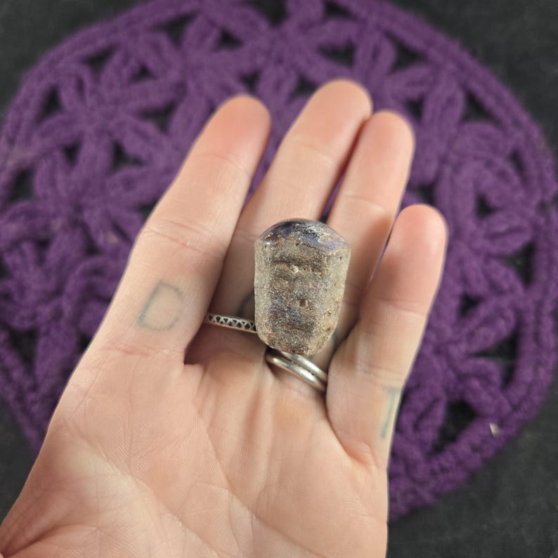 May include: A natural stone ring with a rectangular, textured, light brown body and a dark purple top. The ring is held in a hand with a silver ring on the finger. The background features a purple crocheted pattern.