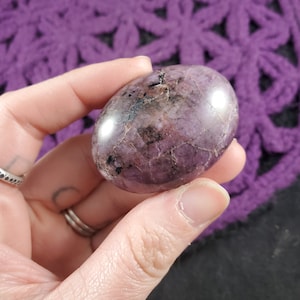 May include: A smooth, polished purple and black gemstone with a natural, irregular shape. The stone is held in a hand, showcasing its size and texture.