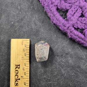 May include: A small, gray, cylindrical object with a dark top, possibly a bottle stopper, next to a wooden ruler. The ruler is marked in inches and has the word "Rocks" printed on it. A purple knitted item is in the background.