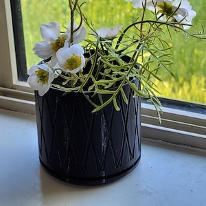 May include: A black planter with a diamond pattern design holds a bouquet of white flowers with yellow centres. The flowers are arranged in a vase on a windowsill.